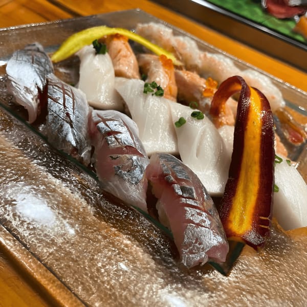 Assorted sushi platter with various fish, shrimp, and a colorful vegetable garnish on a clear plate.